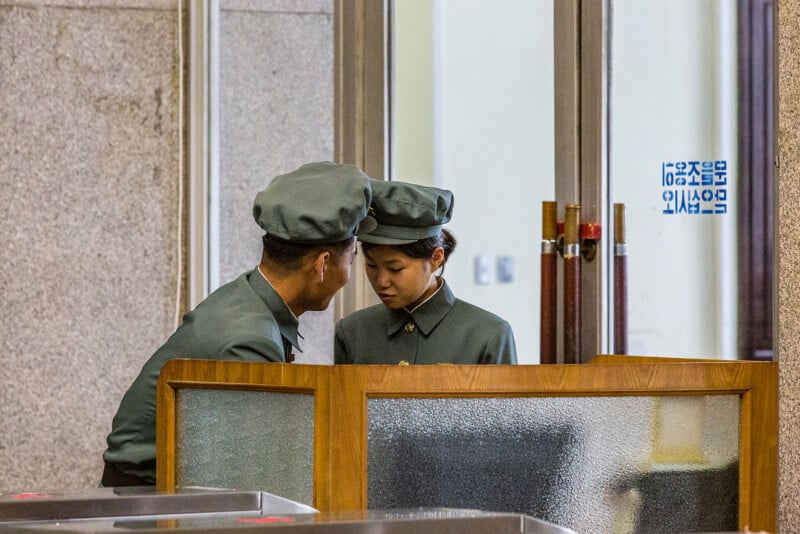 Two individuals in green military uniforms are standing behind a wooden counter, engaged in conversation. Both wear matching green caps and are in a serious discussion. Beside them is a glass door with blue text written in Korean.