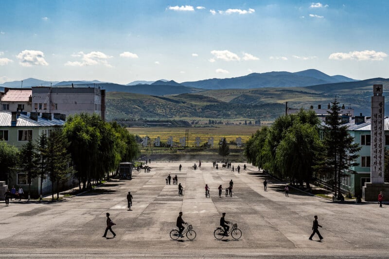 A wide street with a few pedestrians and cyclists is flanked by buildings and trees. In the background, rolling hills and a blue sky with scattered clouds can be seen, creating a serene landscape.