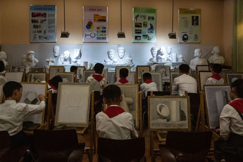 Students in uniform, each sitting at an easel, are sketching various geometric shapes and sculpted busts. The classroom walls feature anatomical charts and educational posters under focused lights.