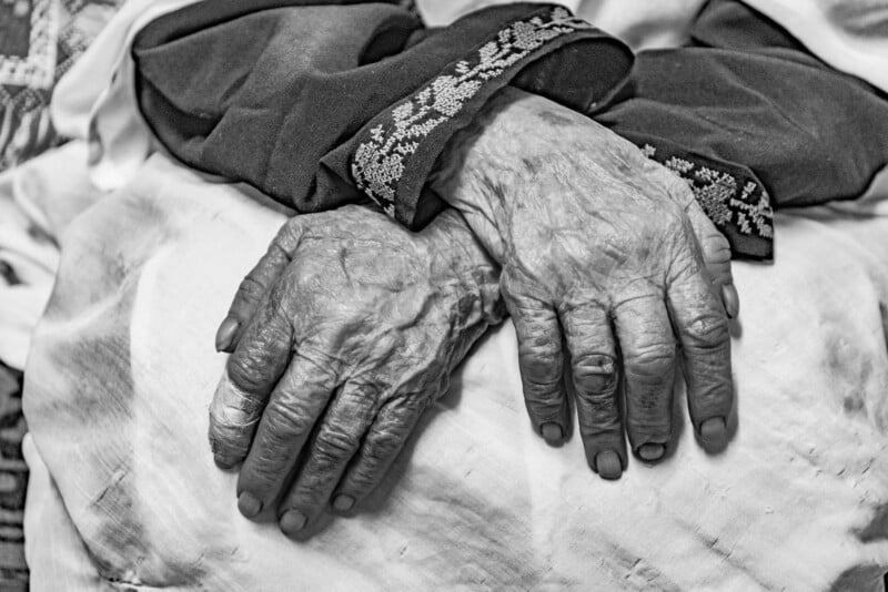 Black and white close-up of an elderly person's hands resting on a light-colored fabric. The hands are adorned with signs of aging, showcasing wrinkles and veins. The individual is wearing a long-sleeved garment with intricate patterns on the cuffs.