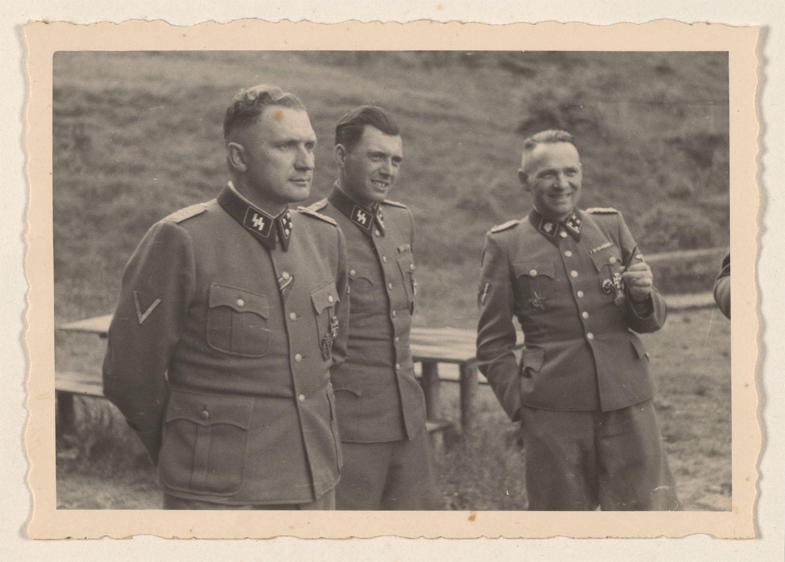 A black-and-white photo shows three men in military uniforms standing outdoors. The man on the left looks ahead with hands behind his back. The middle man and right man, smiling, are engaged in conversation. An outdoor setting with a table is visible in the background.