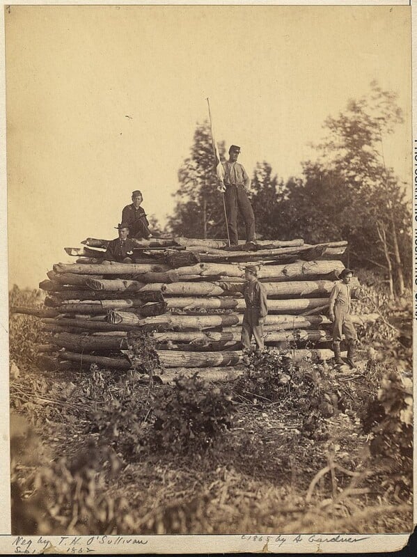 A sepia-toned photograph shows five men standing on and around a large log structure in a wooded area. One man stands on top holding a long pole, while others are positioned at various levels on the logs. The photo is set in a clearing with dense foliage in the background.