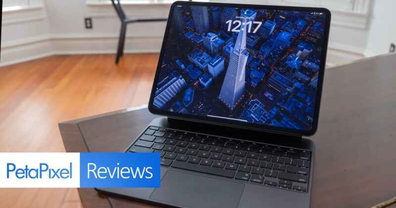 An open iPad Pro on a wooden table displaying a cityscape wallpaper with a digital clock reading 12:17, in a room with wooden floors and white walls, marked with a "PetaPixel Reviews" logo.
