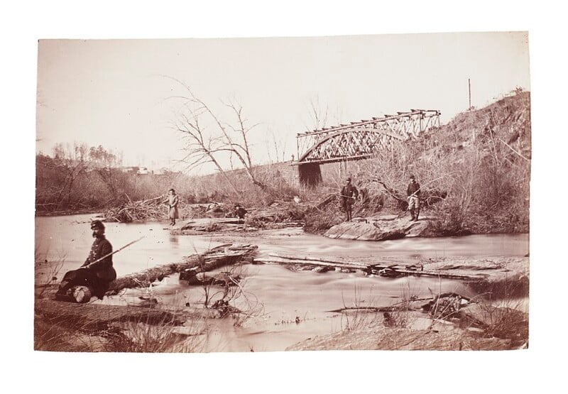 A sepia-toned photograph of five people fishing along the bank of a river. A steel railway bridge spans the river in the background. The surrounding area is filled with bare trees and bushes, indicating a barren landscape.