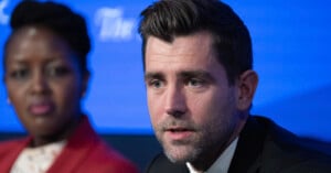 Chris Cox with styled hair and a beard, wearing a black suit, speaks at a panel discussion, with a blurred woman in the background. They are both in front of a blue backdrop with logos.