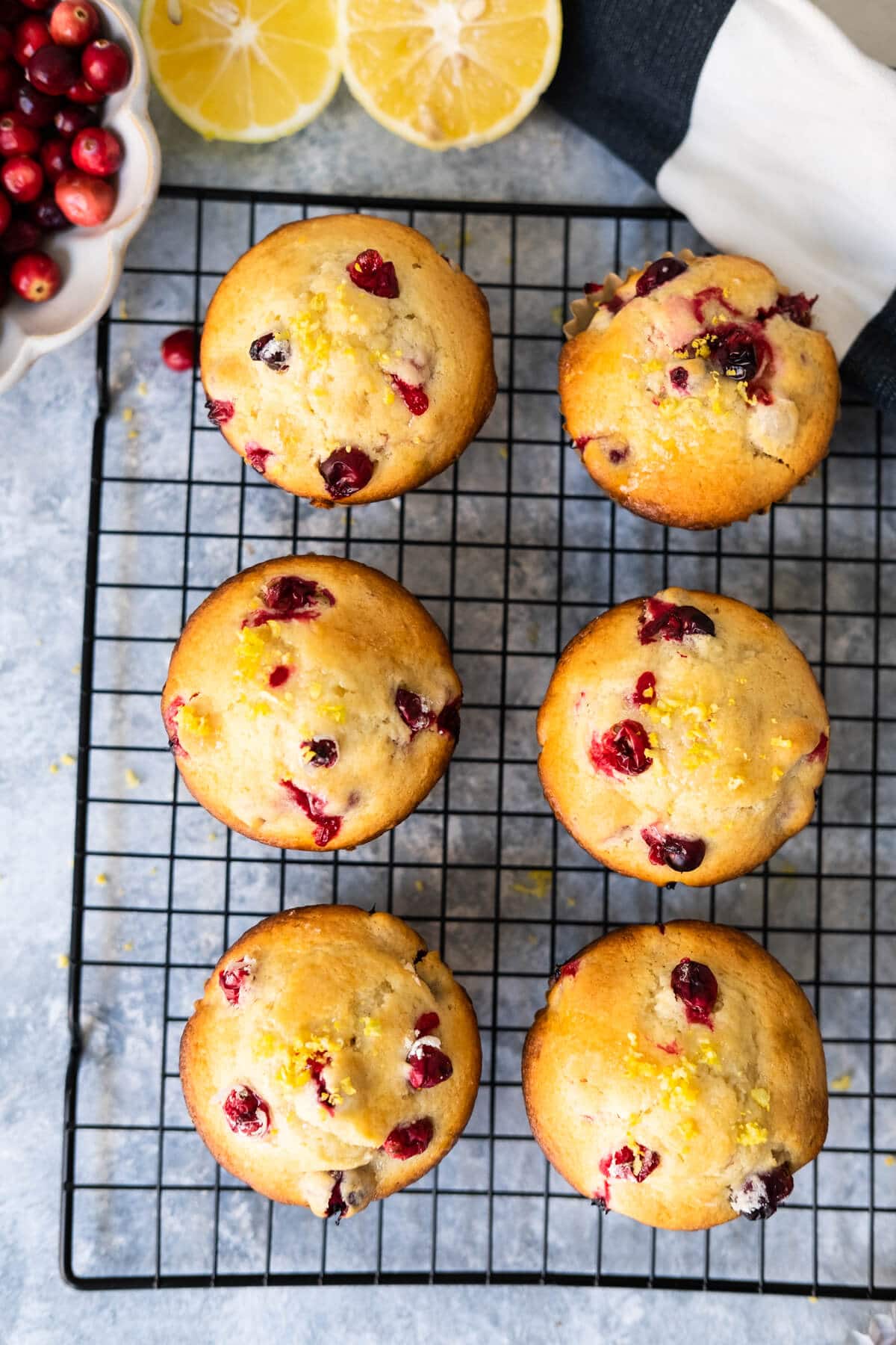 Six cranberry lemon muffins sit on a cooling rack with fresh lemon and cranberries on. the side. 