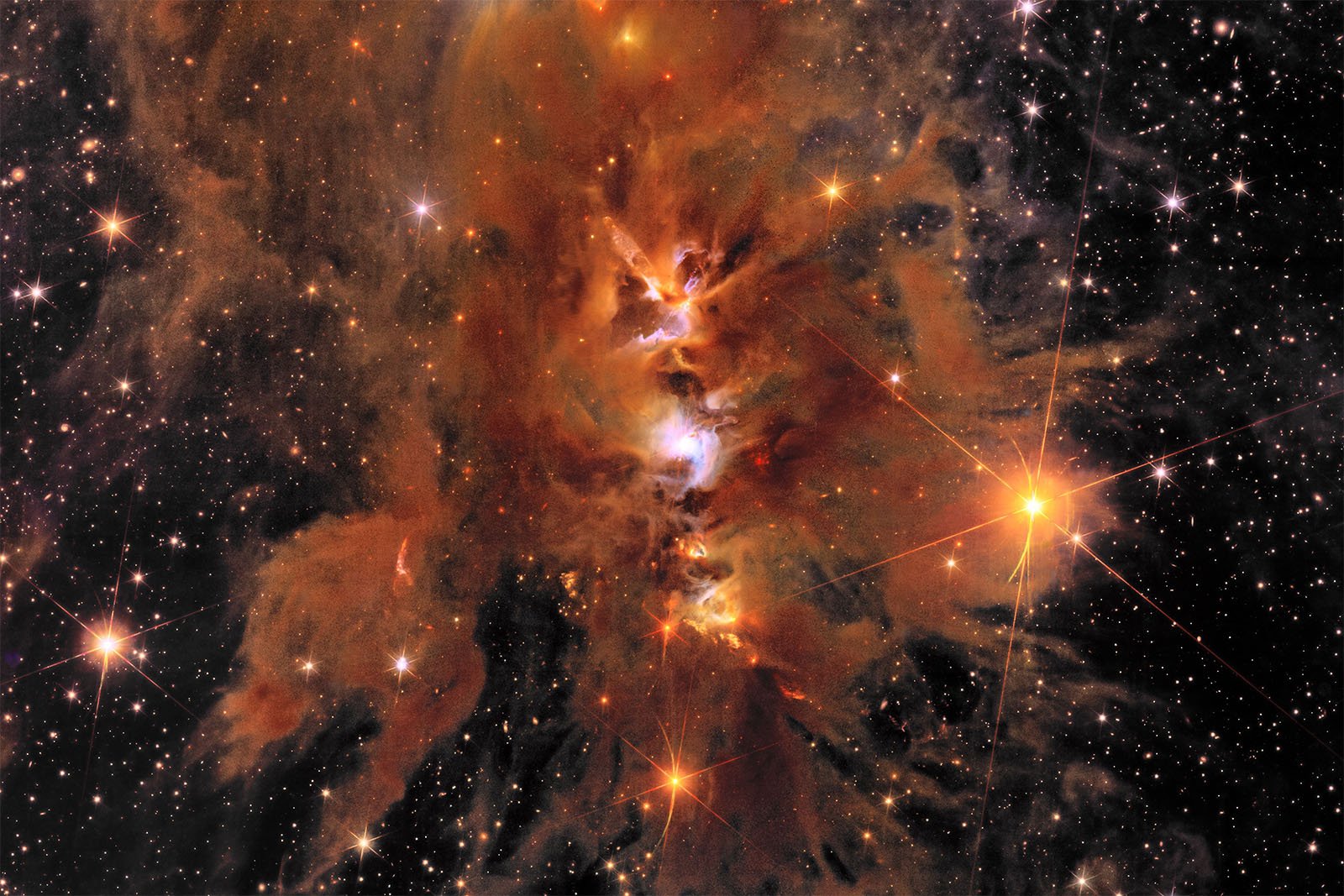 A vibrant, colorful nebula with a mix of orange, red, yellow, and blue hues, surrounded by scattered stars of varying brightness. The nebula has a wispy, cloud-like appearance against the dark backdrop of space. Bright stars shine with light rays extending outward.