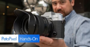 A person wearing a blue shirt holds up a silver and black Fujifilm X-T50 camera with a large lens. In the foreground, the words "PetaPixel Hands-On" are displayed. The background is blurred, focusing attention on the camera.