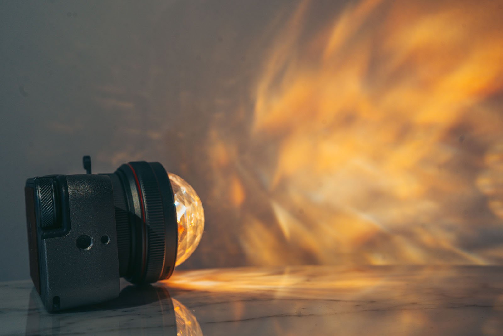 A vintage black camera with a large spherical lens attachment is placed on a marble surface. The lens projects an array of diffused, warm-colored light patterns onto the wall in the background, creating a visually striking effect.
