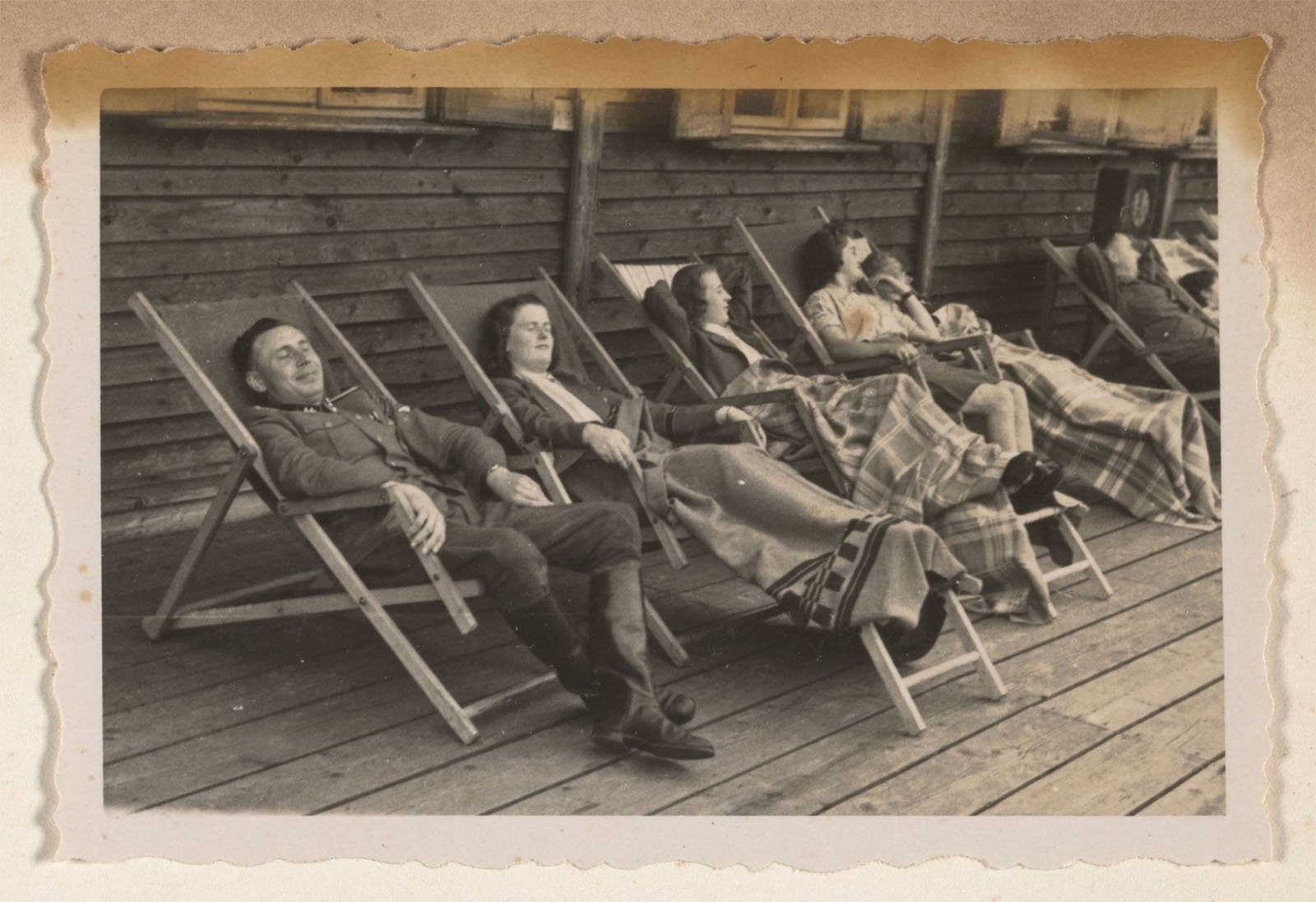 A black-and-white photograph showing four people relaxing on deck chairs arranged in a row on a wooden deck. They are covered with blankets and appear to be napping or resting beside a wooden building, with their arms outstretched holding each other's hands.