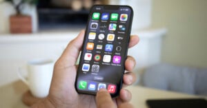 A hand holding an iPhone with a home screen displaying various apps, including Messages, Calendar, Photos, Camera, Maps, Weather, and others. The user's thumb is poised to tap the Mail app icon. A white mug and blurred background elements are visible.