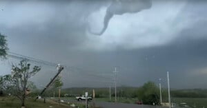 A tornado forms dangerously close to a street, bending utility poles, under a stormy sky. debris is scattered in the air and a white truck is visible in the distance.