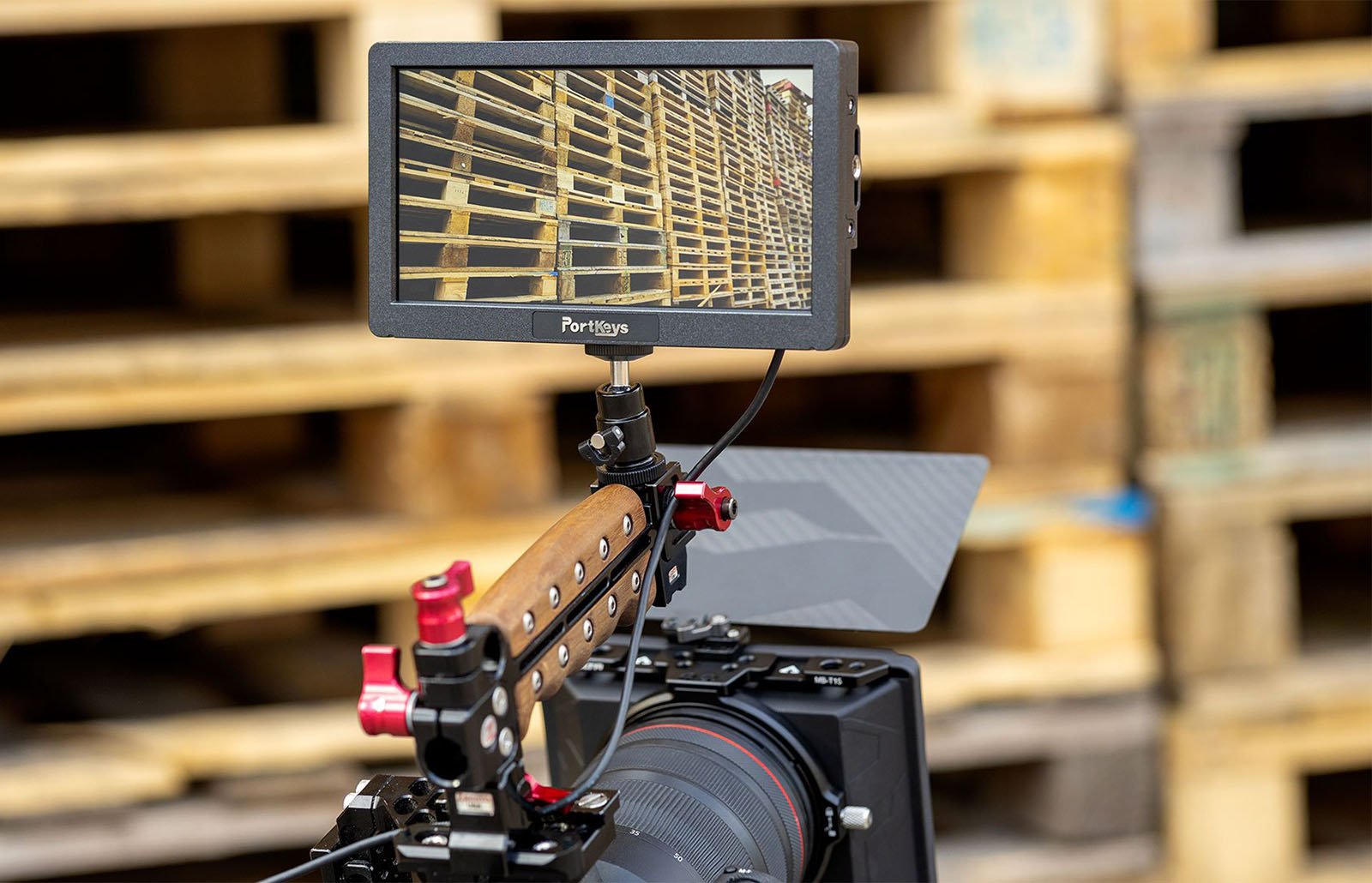 Close-up of a professional camera setup with a wooden handle and attached monitor displaying an image of stacked wooden pallets. The background shows a warehouse setting with more wooden pallets blurred out.