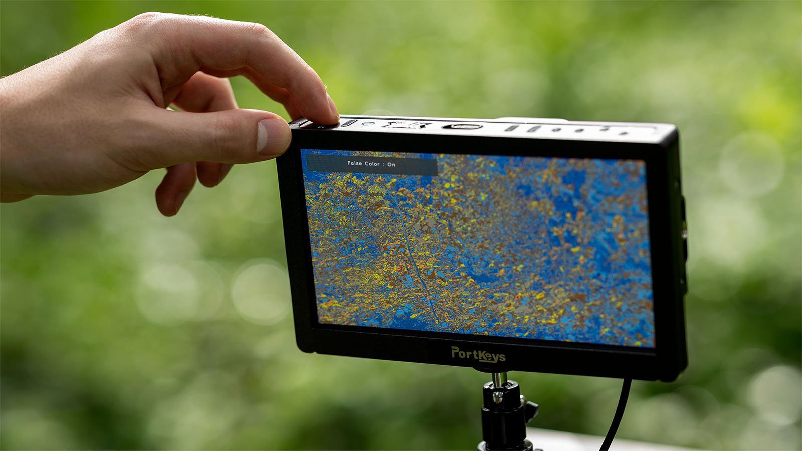 A person adjusts the settings on a PortKeys monitor mounted on a stand. The screen displays an image of a tree with green and yellow leaves against a blue sky. The background is blurred greenery.