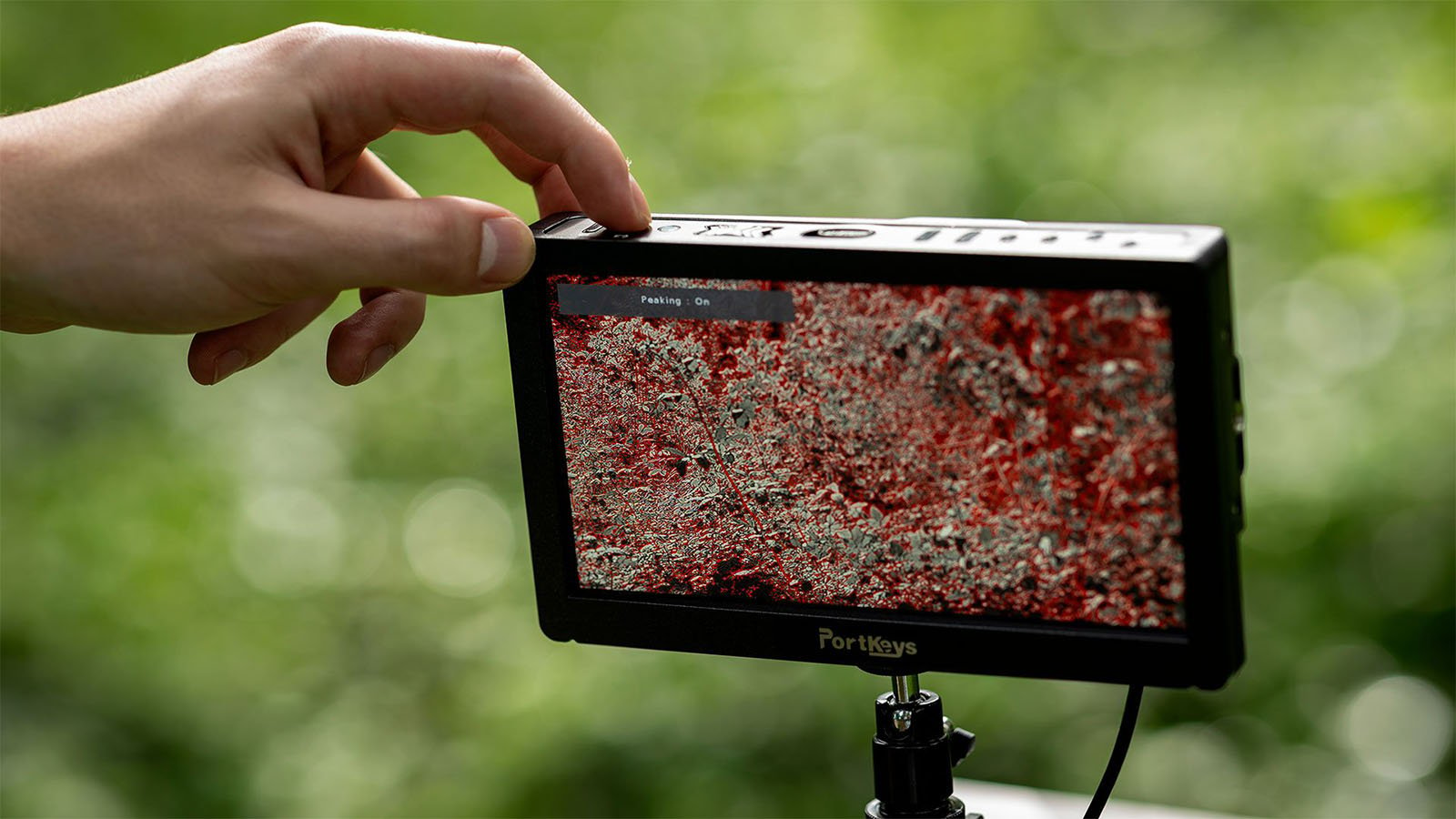 A hand adjusts the settings on a PortKeys camera monitor mounted on a stand. The screen displays a zoomed-in, grainy, red image with the text "Peaking... On" visible. The background is blurred greenery.
