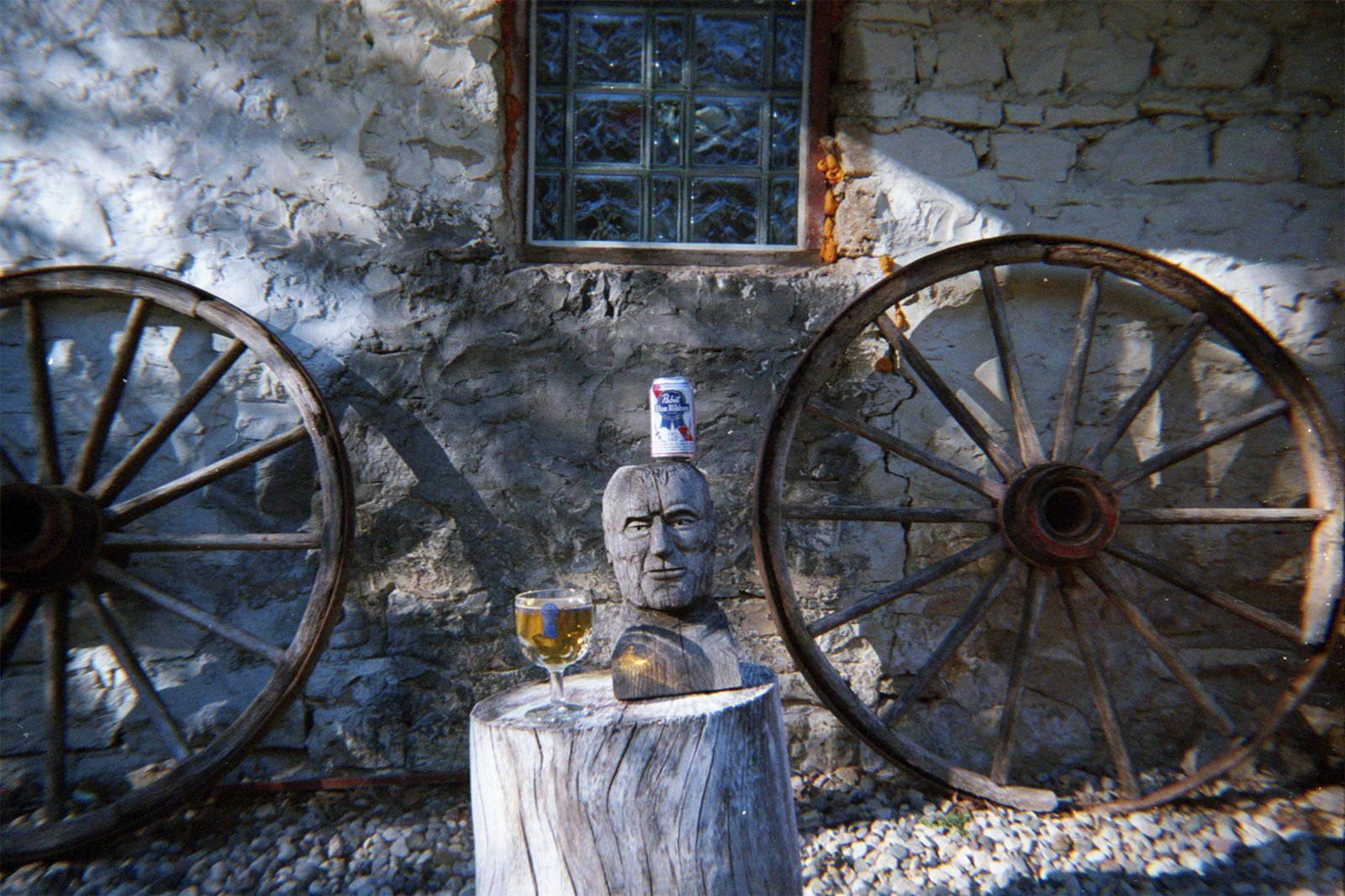 A rustic scene featuring a wooden sculpture of a face sitting on a tree stump. On top of the sculpture is a can of Pabst Blue Ribbon beer. A glass of beer and a baseball glove are also on the stump. Two large wagon wheels lean against a stone wall in the background.