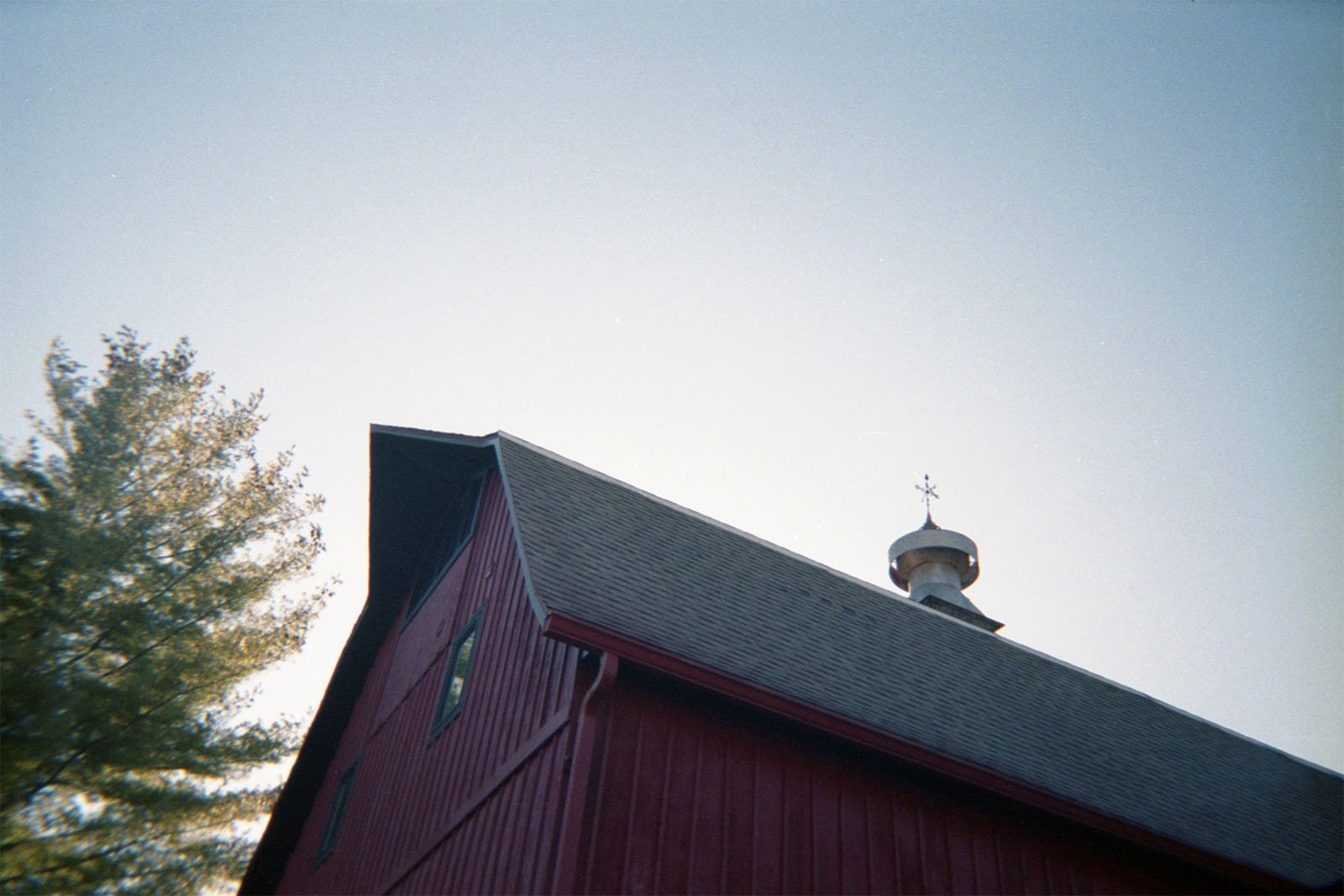 A red barn with a gray shingled roof and a weather vane sits under a clear sky. Part of a tree with green foliage is visible to the left, providing contrast to the barn's red color. The barn has a white cupola on its roof.