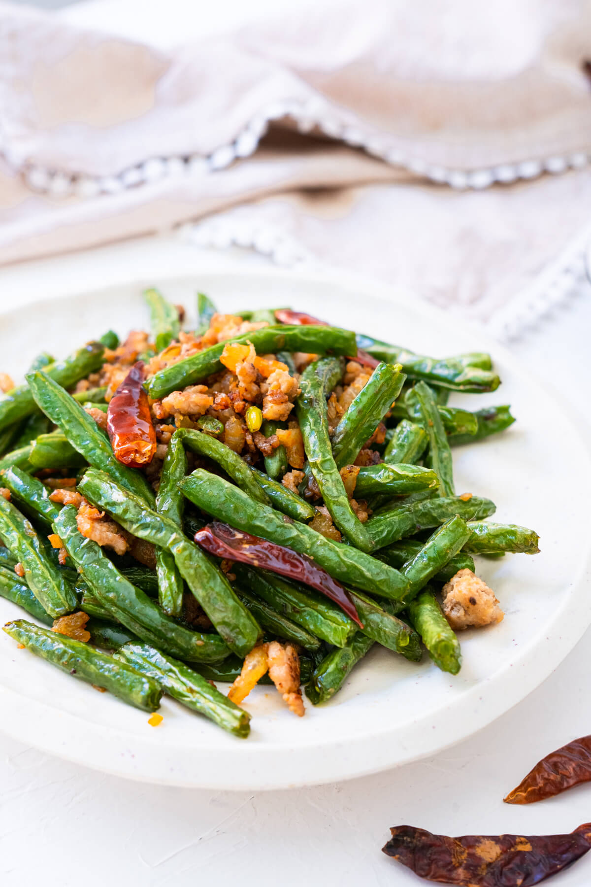 Stir-fried Sichuan green beans in a white platter with a kitchen towel in the back and two pieces of dried chili peppers. 