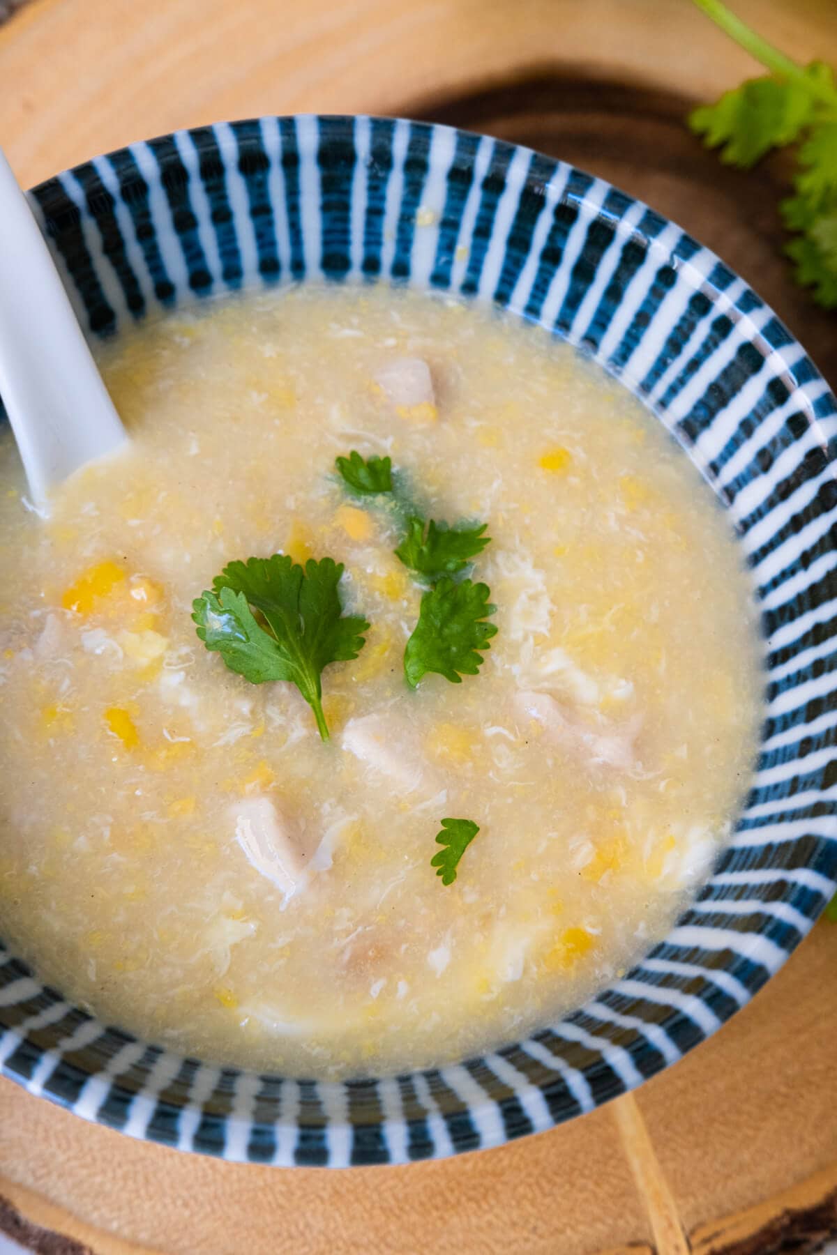 Thick sweet corn and chicken soup in a blue small bowl with spoon, and is served on a wooden plate. 