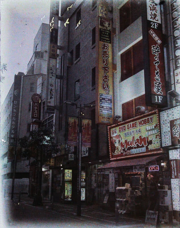 A grainy image of a bustling street in japan at dusk, featuring neon signs, birds flying above, and various storefronts including a dvd and game shop.