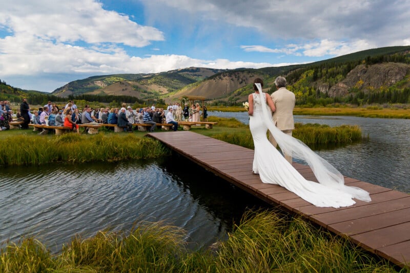 A bride and an older man, possibly her father, walk down a wooden dock toward a gathering of seated guests in an outdoor wedding setting with mountains and a lake in the background. The bride is in a white dress with a long veil, and the venue is surrounded by lush greenery.